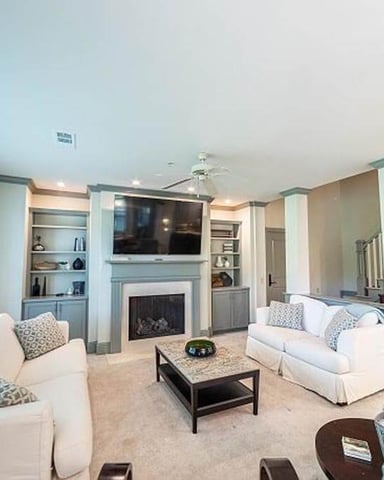 Spacious living room with white upholstered seating, fireplace, wall-mounted TV, built-in shelving, and staircase to upper level