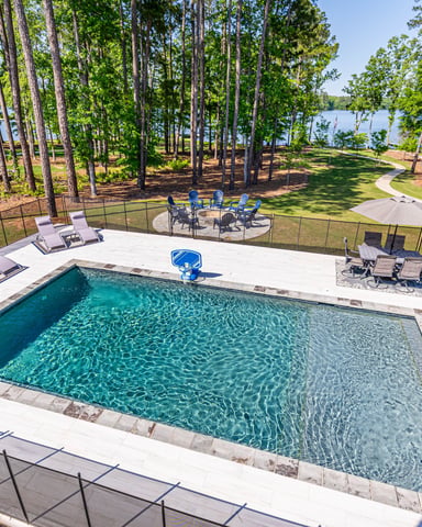 Rectangular swimming pool with lounge chairs and safety fence, surrounded by tall pine trees and lake view beyond