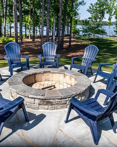 Stone fire pit surrounded by blue Adirondack chairs on patio, with lake and tall trees in background