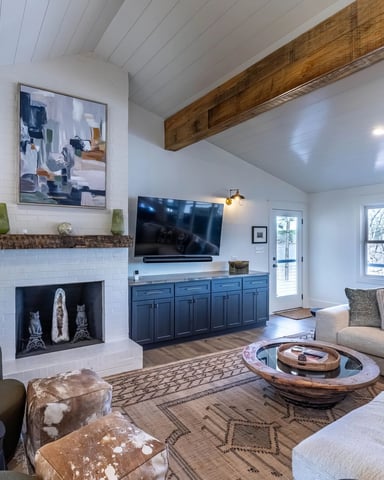 Modern living room with white shiplap ceiling, exposed wood beam, fireplace with TV, blue cabinetry, and lake views through large windows