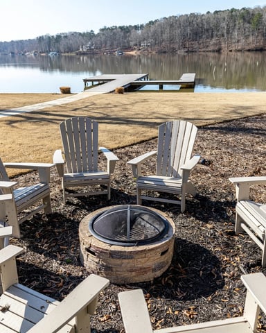 Adirondack chairs arranged around fire pit on sandy beach overlooking calm lake with forested shoreline
