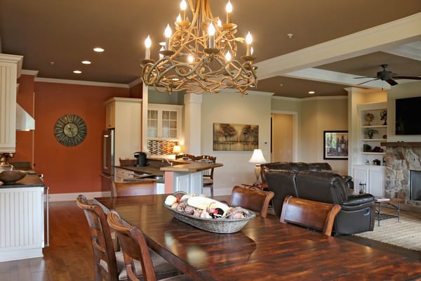 Open-plan living space with dining table, kitchen, and seating area, ornate gold chandelier overhead, stone fireplace visible in background.