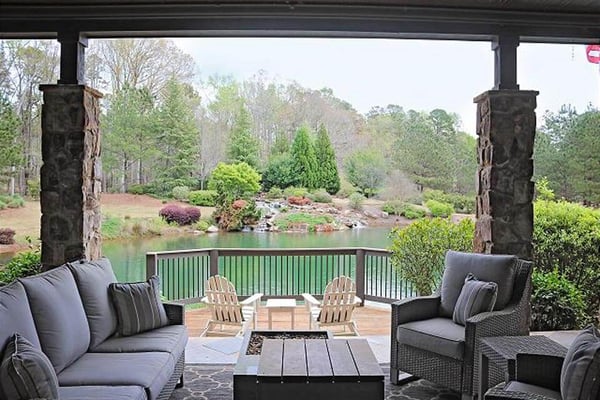 Covered patio with gray seating, fire pit, and Adirondack chairs overlooking pond and wooded landscape