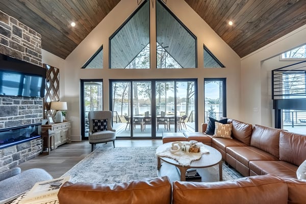 Modern living room with vaulted wood ceiling, large windows overlooking lake, brown leather sectional, and stone fireplace with TV.
