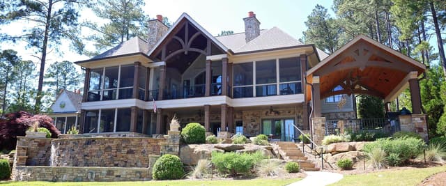 Two-story stone and wood house with screened porches, stone chimney, and covered pavilion surrounded by pine trees and manicured landscaping.