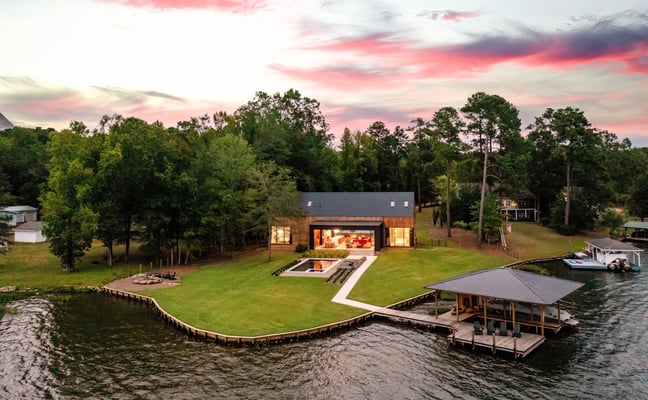Modern waterfront home with lit interior, dock, and boathouse at sunset surrounded by trees